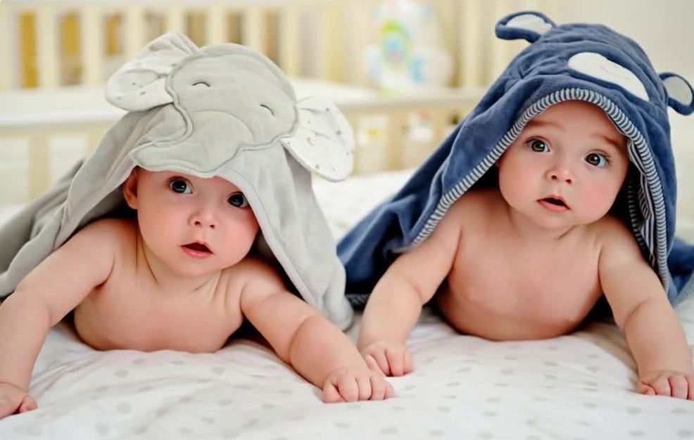 Two infants lying on a bed, propped on their forearms, wearing hooded towels.