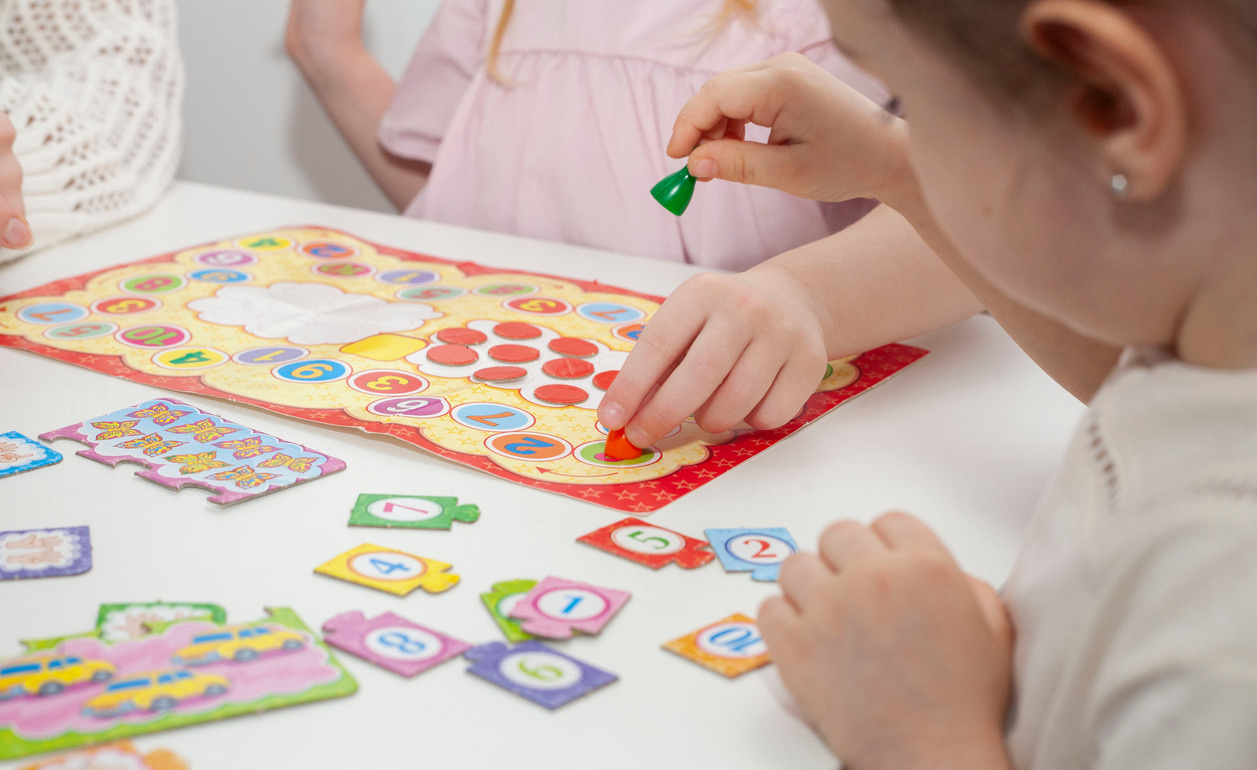A child's hands placing a coloured counter onto a numbered board game at a white table.