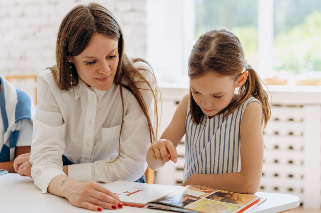 A tutor and a child reading together at a desk by a window