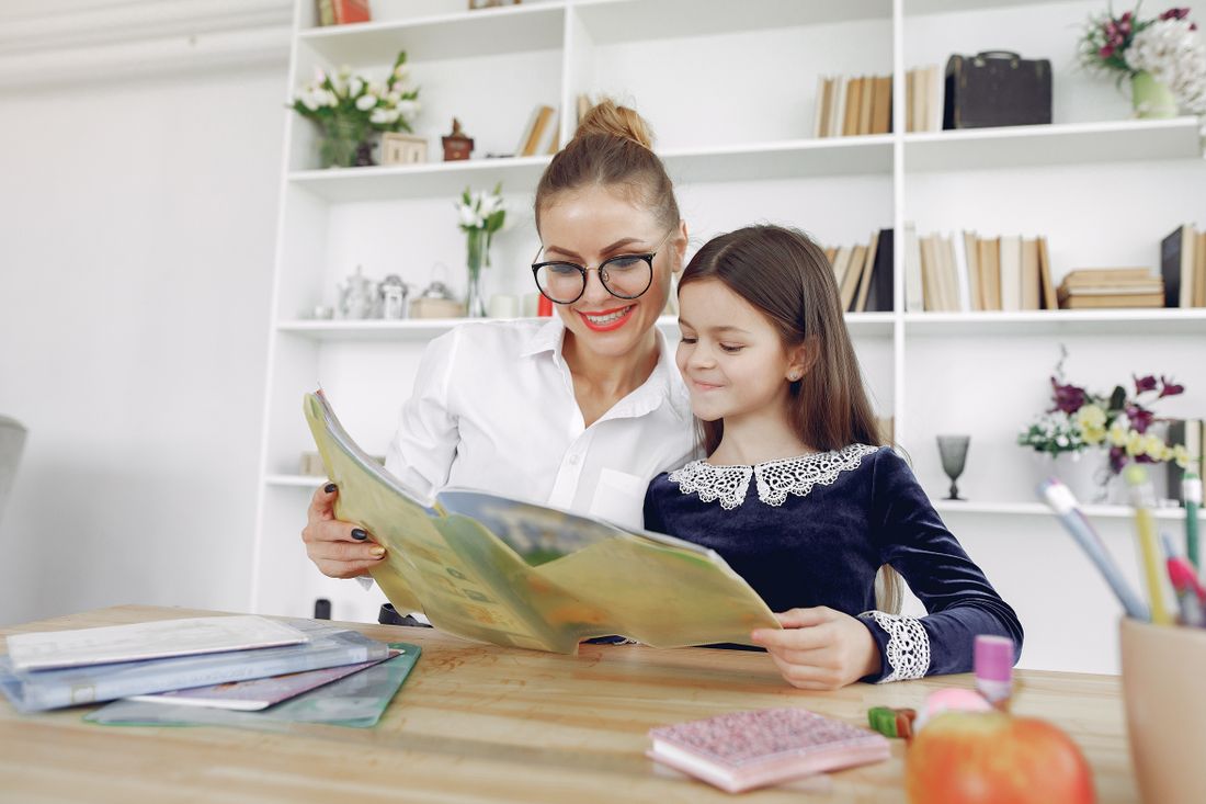 A tutor and a child reading a book together beside a bookshelf